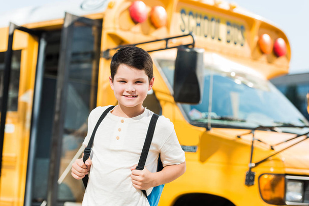 smiling little schoolboy with backpack standing in front of school bus - Zdjęcie, obraz
