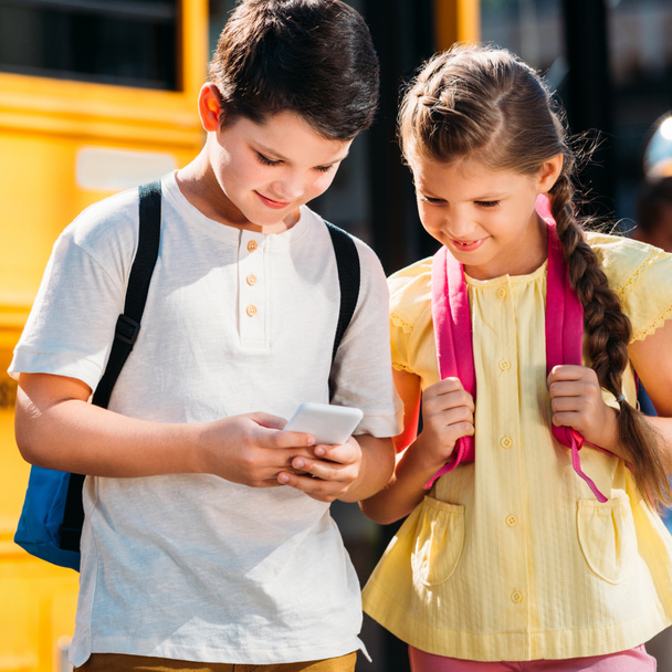 happy little scholars using smartphone together in front of school bus - 写真・画像