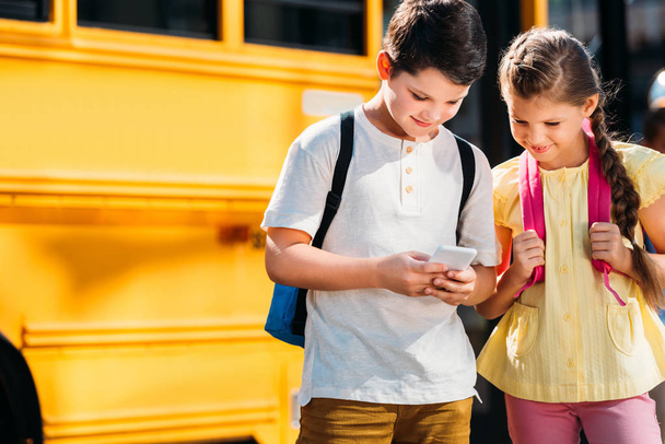 愛らしい小さな学者一緒に学校のバスの前でスマート フォンを使用して - 写真・画像