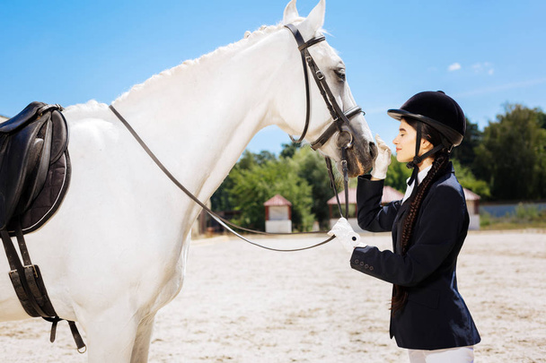 Beautiful slim woman loving equestrianism looking at white horse - Photo, Image