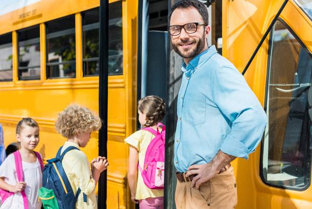 male teacher looking at camera while pupils entering school bus blurred on background - Photo, Image