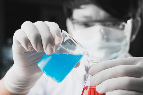 Chemist student boy pouring blue liquid in beger glass to erlenmeyer flask chemistry with red liquid on biochemical experiment. Mix water in laboratory. - Photo, image