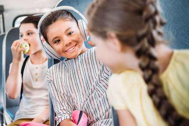 mooie kleine schoolmeisje luisteren muziek met koptelefoon tijdens het rijden op de bus van de school met klasgenoten - Foto, afbeelding