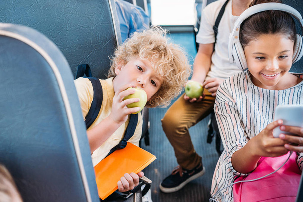 adorable little schoolboy eating apple while riding on school bus with classmates - Fotoğraf, Görsel