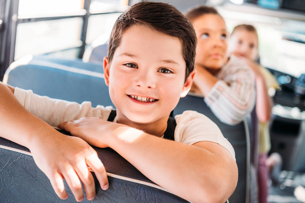 close-up portrait of smiling little schoolboy riding on school bus with blurred classmates on background - 写真・画像