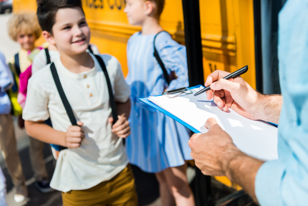 vágott lövés a tanár írásban listát a vágólapra, míg a tanulók belépő iskola busz homályos a háttér - Fotó, kép