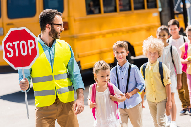 красивый дорожный охранник пересекает дорогу со школьниками перед школьным автобусом
 - Фото, изображение