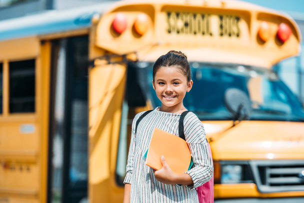 happy little schoolgirl with notebooks looking at camera in front of school bus - Fotografie, Obrázek
