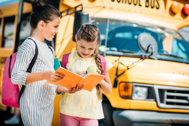 adorable little schoolgirls with notebook discussing homework in front of school bus - Photo, image