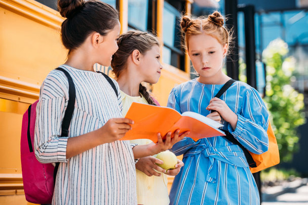 group of little schoolgirls with notebook talking in front of school bus - Фото, изображение