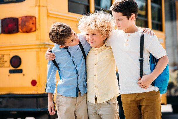 ευτυχής λίγο σχολιαρόπαιδα Αγκαλιάζοντας και μιλώντας μπροστά από το σχολικό λεωφορείο - Φωτογραφία, εικόνα