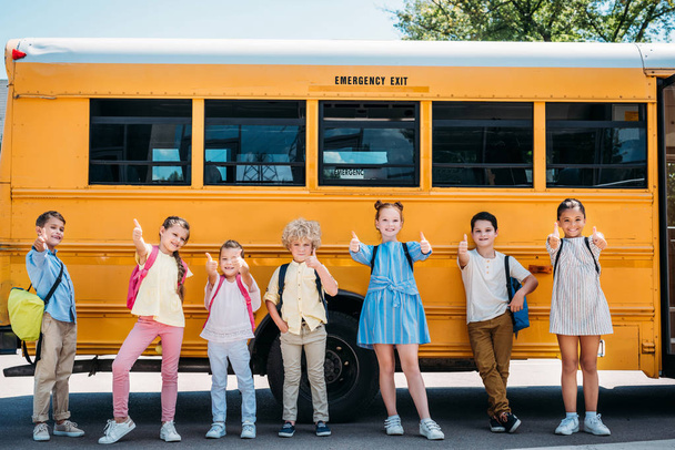 group of adorable schoolchildren standing in front of school bus and showing thumbs up - Foto, afbeelding