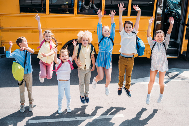group of adorable schoolchildren jumping in front of school bus and looking at camera - Foto, afbeelding