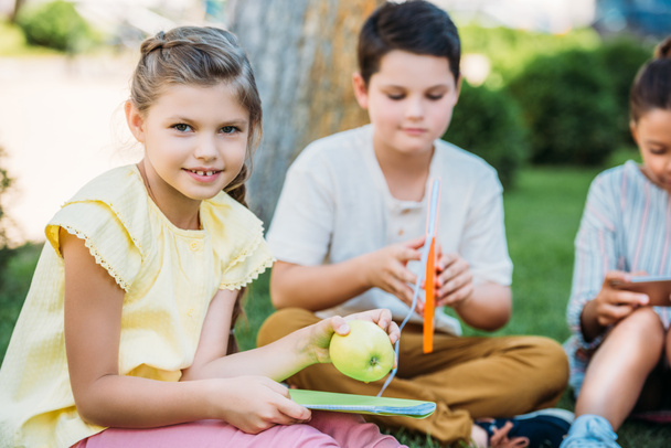 adorable little schoolgirl with apple and book sitting on grass with classmates - Photo, Image