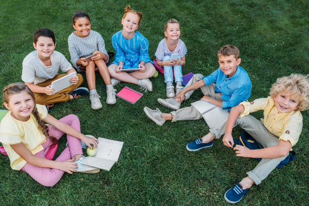hoge hoekmening van groep gelukkig scholieren zittend op het gras met boeken en apparaten - Foto, afbeelding