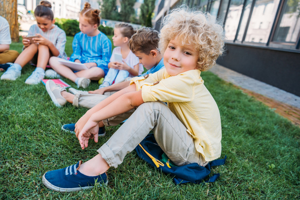 adorable curly schoolboy sitting on grass with classmates - Photo, image