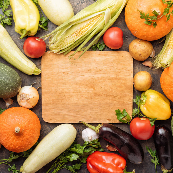 Assortment of fresh vegetables on dark old metal pan background and chopping wooden board. Courgettes, corn, pumpkin, zucchini, potato, pepper, onions, garlic - Foto, Bild