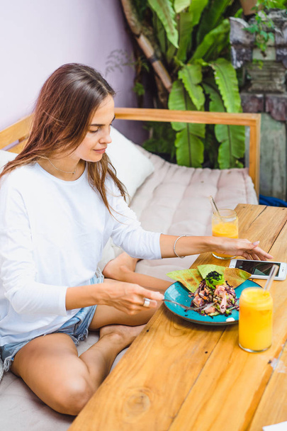 Uma bela mulher toma café da manhã em um café elegante, um café da manhã saudável, frutas, suco espremido na hora, localização tropical
. - Foto, Imagem