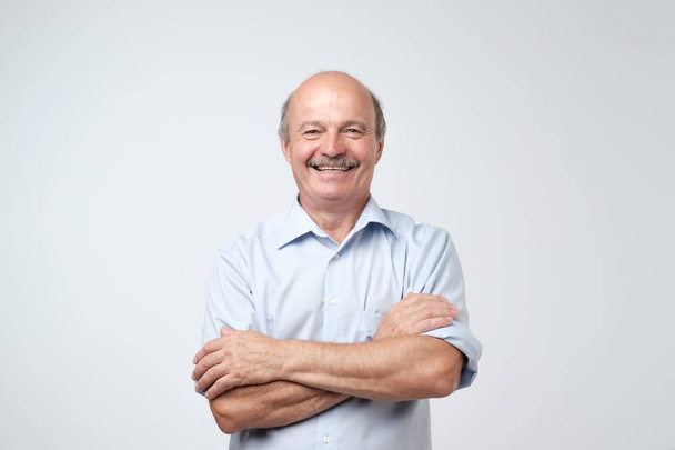 Okouzlující hezký starší muž v neformálním modré tričko držet rukama zkříženýma a usmívá se přitom stát izolovaných na bílém pozadí. - Fotografie, Obrázek