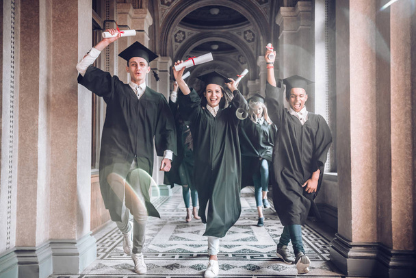 Celebrating their achievements together.University was the best years of their lives!Group of smiling university students holding their diplomas and running after being graduated in university hall. - Фото, зображення
