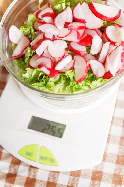 Salada dietética com rabanete e alface em uma tigela de vidro em pé nas balanças da cozinha. O conceito de alimentação saudável adequada. Contagem de calorias e produtos de perda de peso
 - Foto, Imagem