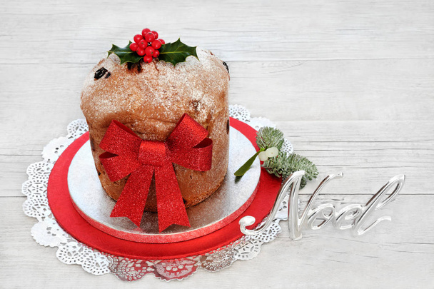 Традиционный рождественский торт из панеттона с серебряным знаком Ноэля, красным блестящим бантом и зимним холли на деревенском дубовом фоне. Итальянский хлеб
. - Фото, изображение