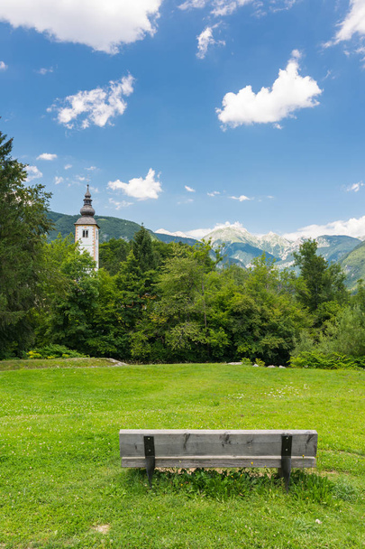 Bohinj church in the Ribcev Laz village, Slovenia. View of old ancient church from the park with bench. Slovenia alps with Triglav in background, grass and park in foreground - Photo, Image
