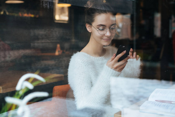 smiling young girl freelancer with glasses resting in a cafe, looking into her smartphone - 写真・画像