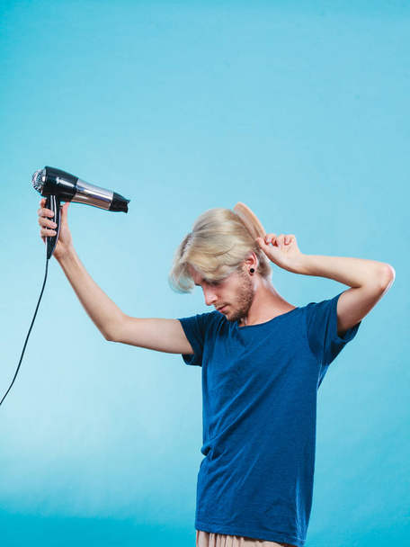 Style and fashion. Young trendy male hairstylist barber with new idea of look changing. Blonde man holding hair dryer and comb creating new hairdo, on blue - Foto, immagini