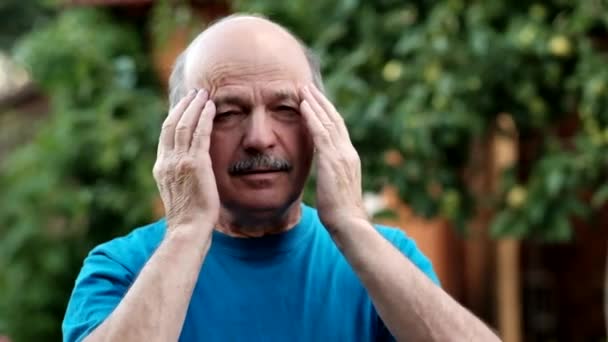 Homem caucasiano mostrando o quanto sua cabeça dói, parecendo miserável e exausto de pé ao ar livre no jardim
 - Filmagem, Vídeo