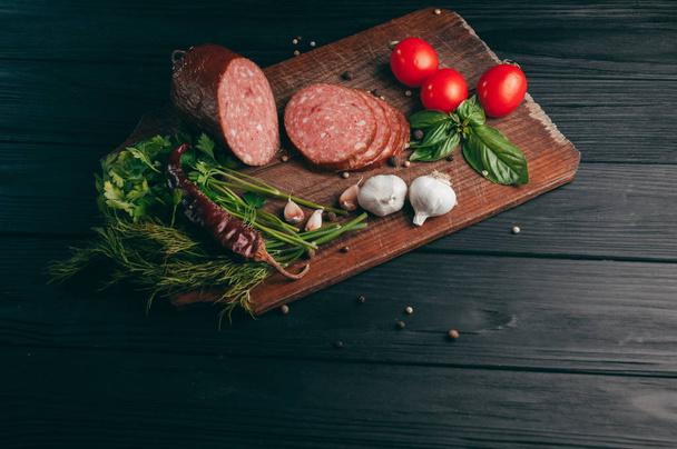 Smoked salami on a black table and a brown board. Sausages with rosemary, garlic and pepper and tomato cherry, dill, parsley - Foto, immagini