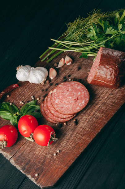 Smoked salami on a black table and a brown board. Sausages with rosemary, garlic and pepper and tomato cherry, dill, parsley - Foto, Imagem