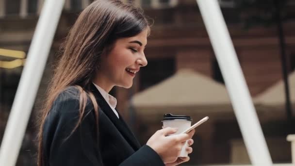 Professional young businesswoman walking on urban street using smartphone and drink coffee. Concept: new business, communication, banker. Outside, slow motion - Πλάνα, βίντεο