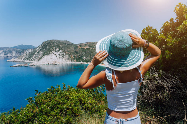 Petani beach Kefalonia. Young woman holding blue sun hat enjoying beautiful panorama of blue bay lagoon surrounded by steep cliff coastline. Greece - Foto, Imagen
