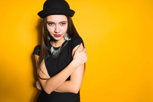 luxurious attractive girl in a fashionable black hat posing on a yellow background, in the ears expensive earrings - Foto, afbeelding