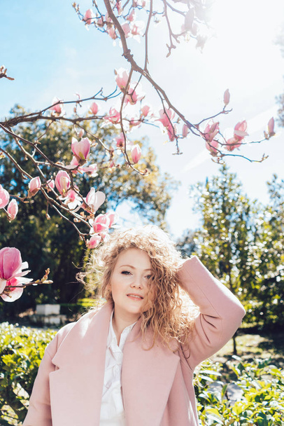 luxurious curly woman posing in the sun in the park, against the background of a blooming magnolia, smiling - Фото, изображение
