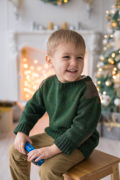 little boy makes funny faces on the background of the fireplace with New Year decor, garlands, Christmas trees, gifts. Merry Christmas - Фото, зображення