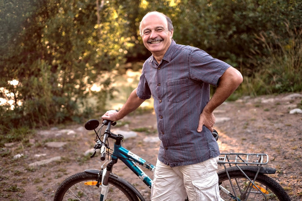 Joyful senior man standing with a bike in a park on a beautiful sunny day. Active life in senior age. Concept of sport on vacation - Photo, Image