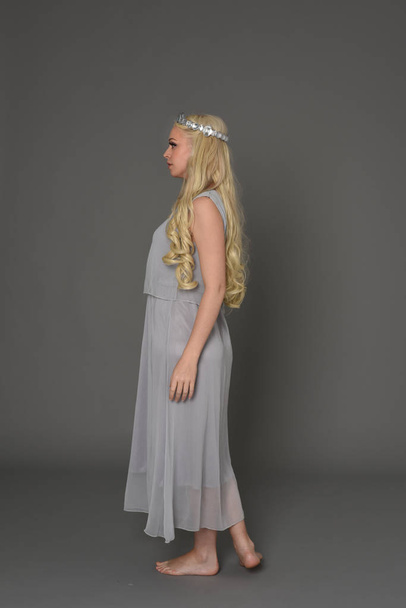 full length portrait of blonde girl wearing crow and grey dress, standing pose in side profile.  grey studio background. - Photo, Image