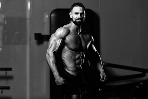 Hombre joven de pie fuerte en el gimnasio y músculos flexionantes - Muscular atlético culturista modelo de fitness posando después de los ejercicios - Foto, imagen