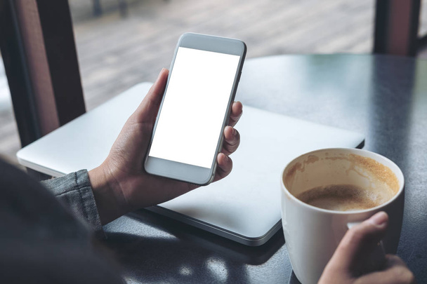 Mockup afbeelding van mobiele telefoon met leeg wit scherm met laptop; deze keer en koffie cup op houten tafel in café - Foto, afbeelding