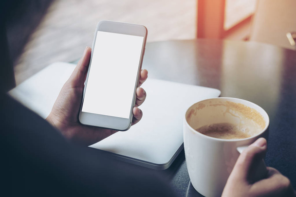 Изображение деловой женщины, держащей в руках мобильный телефон с чистым белым экраном и кофейной чашкой на деревянном столе в кафе
 - Фото, изображение