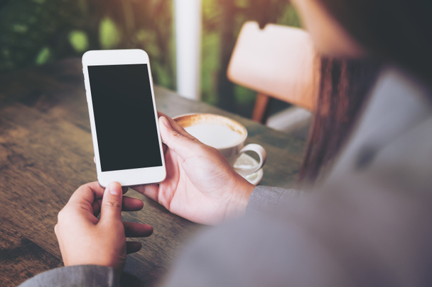 Изображение рук, держащих белый мобильный телефон с чистым белым экраном с чашкой горячего кофе на деревянном столе в кафе
 - Фото, изображение