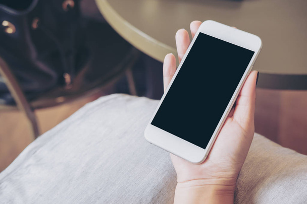 Imagem Mockup de mãos segurando telefone celular branco com tela preta em branco no travesseiro com fundo de piso de madeira
 - Foto, Imagem