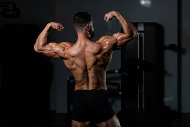 Young Man Standing Strong In The Gym And Flexing Muscles - Muscular Athletic Bodybuilder Fitness Model Posing After Exercises - 写真・画像
