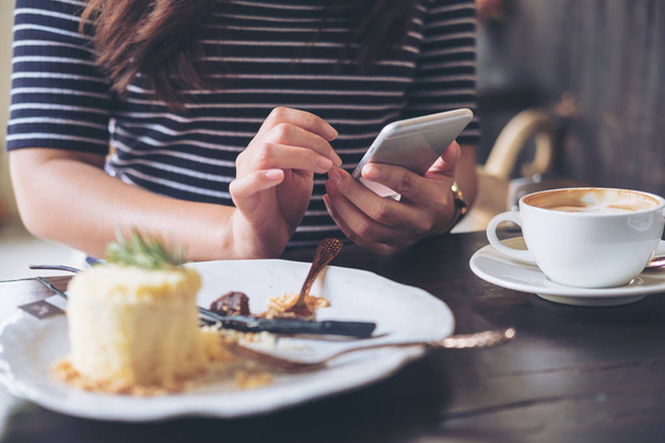 A woman using smart phone with cake and coffee cup on wooden table in restaurant  - Foto, Bild