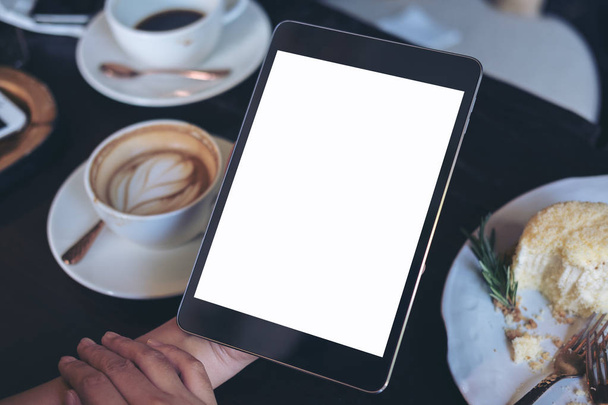 Mock up imagem de mãos de mulher de negócios segurando computador tablet preto com tela branca em branco, bolo e xícaras de café na mesa de madeira no café
  - Foto, Imagem