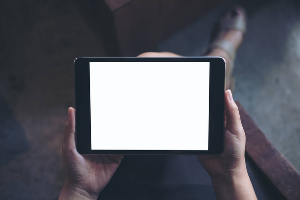 Imagen burlona de la mano de la mujer sosteniendo PC tableta negra con pantalla blanca en blanco en el muslo con fondo de madera en la cafetería moderna
 - Foto, Imagen