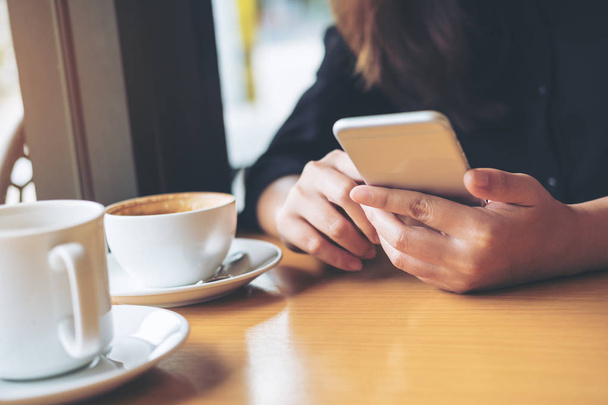 eine Frau mit Smiley-Gesicht hält Smartphone mit Kaffeetasse auf Holztisch im Café - Foto, Bild