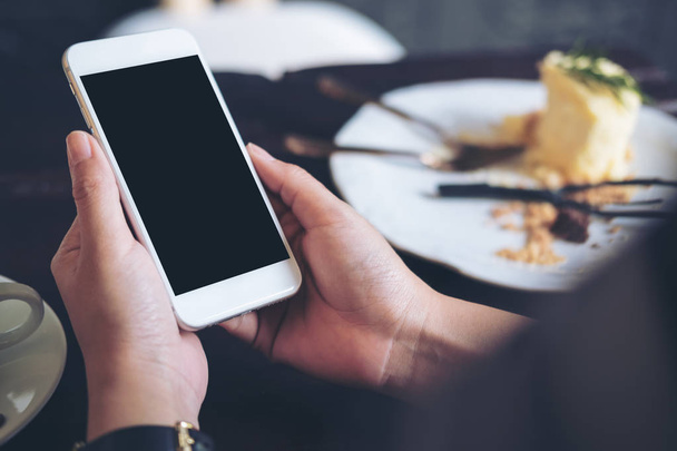 Mockup image of hands holding white mobile phone with blank black screen with a plate of cake on wooden table in restaurant - Foto, immagini